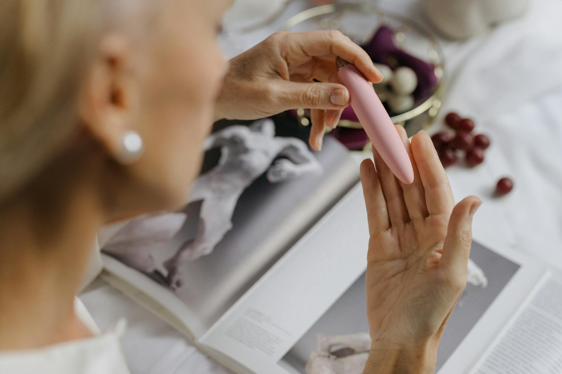 Close-up of hands holding a pink vibrator with art books nearby, representing intimate reconnection and self-discovery.